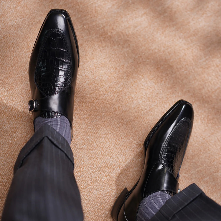 A pair of men's black leather dress shoes with crocodile-embossed panels on the toes, styled with dark pinstripe trousers and gray striped socks on a beige carpeted floor.