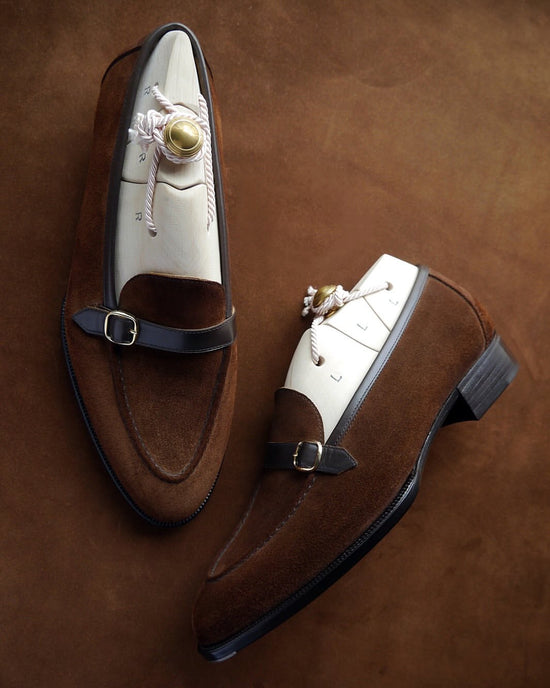 A pair of brown suede and white leather mule loafers with dark brown straps and gold buckle details, arranged on a textured brown background.