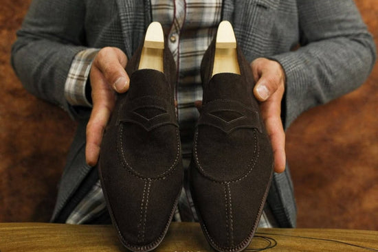 Brown suede loafers held by a person wearing a gray coat with plaid lining.