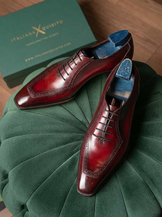 A pair of glossy dark red-brown leather dress shoes with brogue detailing, displayed on a green velvet ottoman with the brand's green box visible in the background.