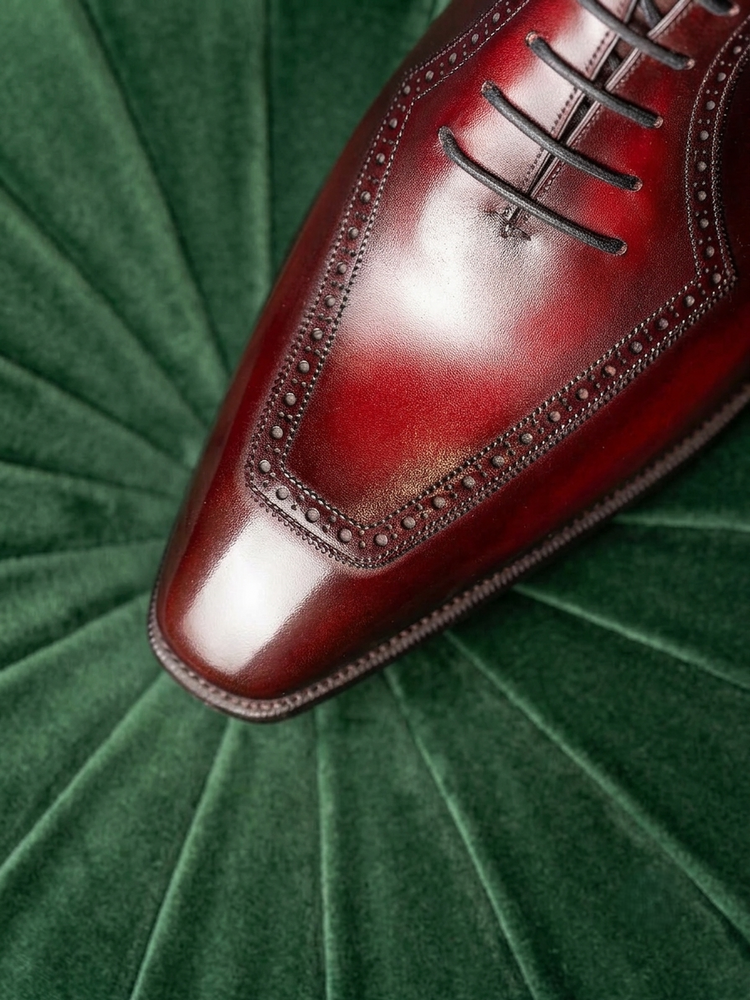 A close-up of a polished dark red oxford shoe toe with decorative brogue detailing, resting on a textured green velvet background.