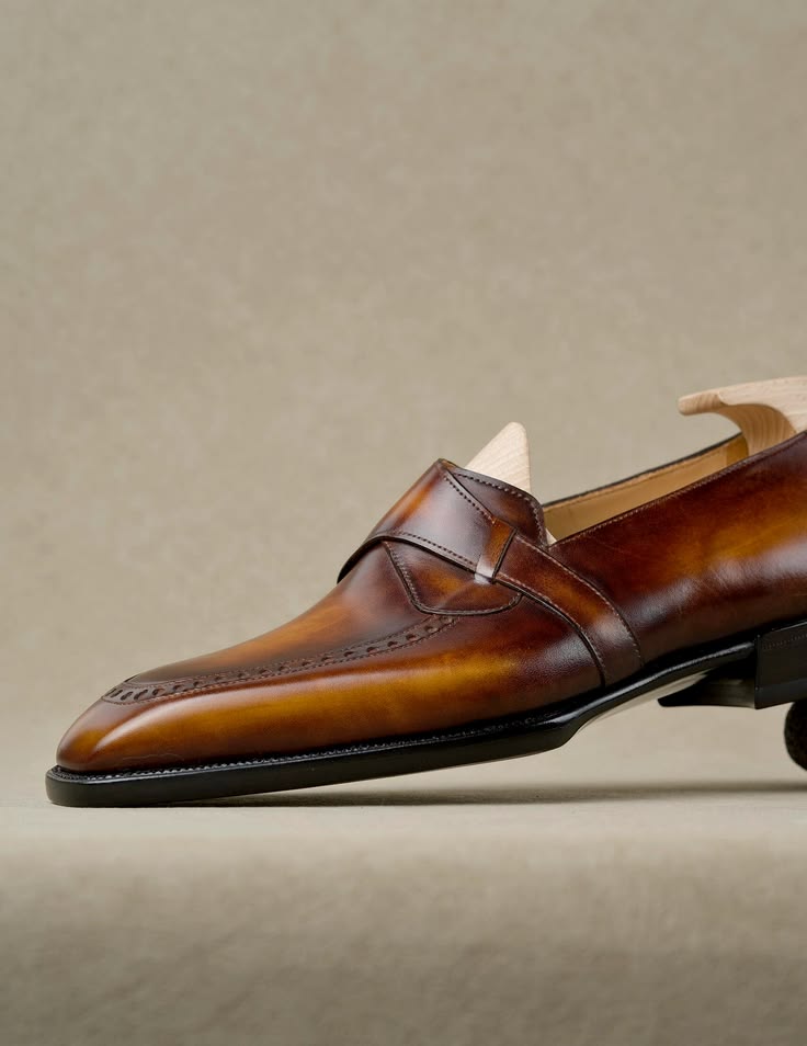A close-up side view of a brown leather penny loafer with brogue detailing, shown on a light beige textured background.