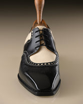 A men's wingtip oxford shoe in black and cream leather, shown from a front-facing perspective on a neutral beige background.