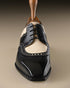 A men's wingtip oxford shoe in black and cream leather, shown from a front-facing perspective on a neutral beige background.