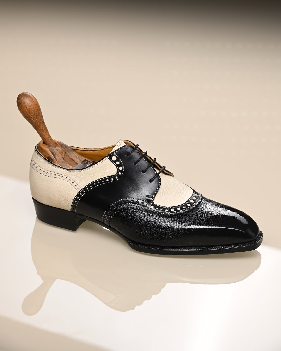 A men's wingtip oxford shoe in black and cream leather, displayed on a light beige background with a wooden shoe tree inserted.
