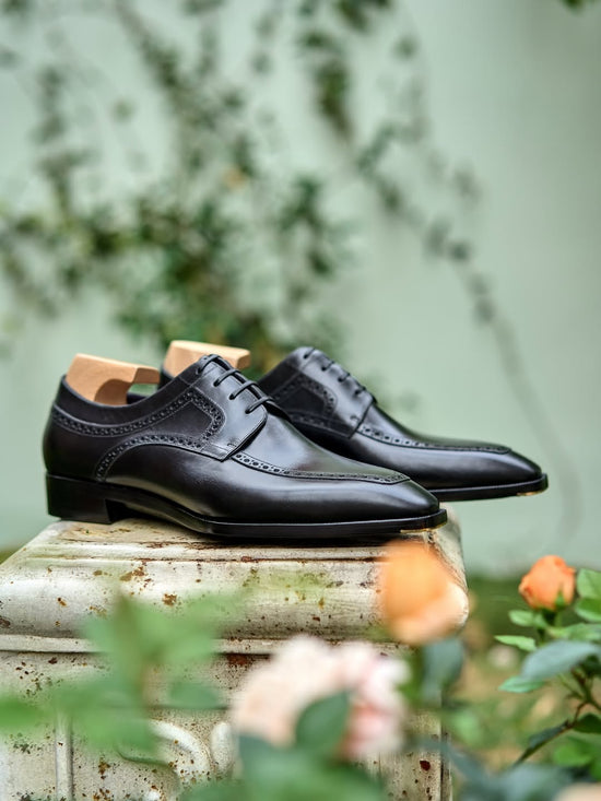 A pair of black leather oxford dress shoes with brogue detailing, displayed on a weathered stone pedestal with blurred green foliage and peach flowers in the background.