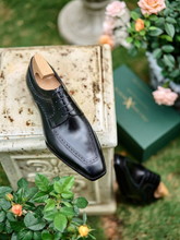 A black leather dress shoe with brogue detailing and a wooden shoe tree insert, displayed on a weathered stone pedestal outdoors with roses, a green box, and another shoe in the background.