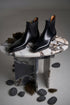 A pair of black leather Chelsea boots with a glossy finish, displayed on a round marble-topped table against a neutral gray background.