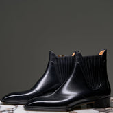 A pair of black leather Chelsea boots with elastic side panels, displayed on a round marble-topped pedestal against a dark gray studio background.