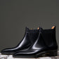 A pair of black leather Chelsea boots with elastic side panels, displayed on a round marble-topped pedestal against a dark gray studio background.