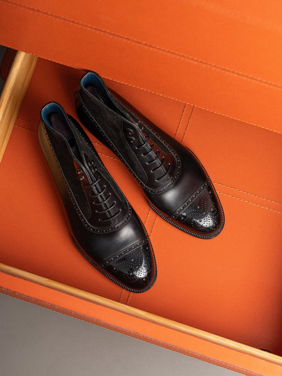A pair of black leather cap-toe brogue oxford shoes with decorative perforated detailing on the toe, displayed inside an orange textured box.