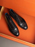 A pair of black leather cap-toe brogue oxford shoes with decorative perforated detailing on the toe, displayed inside an orange textured box.