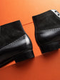 Close-up of a pair of black leather and suede ankle boots with brogue detailing, positioned diagonally on a vibrant orange background.