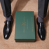 A pair of black embossed leather monk-style dress shoes worn with dark pinstriped trousers and gray socks, arranged symmetrically around a dark green box printed with the brand name 'ITALIANO LOUISITO'.