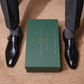 A pair of black embossed leather monk-style dress shoes worn with dark pinstriped trousers and gray socks, arranged symmetrically around a dark green box printed with the brand name 'ITALIANO LOUISITO'.