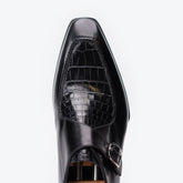 A close-up top-down view of a black leather dress shoe with a crocodile-embossed panel on the vamp and a silver buckle strap, set against a plain white background.