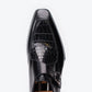 A close-up top-down view of a black leather dress shoe with a crocodile-embossed panel on the vamp and a silver buckle strap, set against a plain white background.