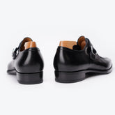 A pair of black leather double monk-style dress shoes shown from the rear, featuring a polished finish, black soles, and light brown wooden shoe trees inside, set against a clean white background.