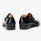 A pair of black leather double monk-style dress shoes shown from the rear, featuring a polished finish, black soles, and light brown wooden shoe trees inside, set against a clean white background.