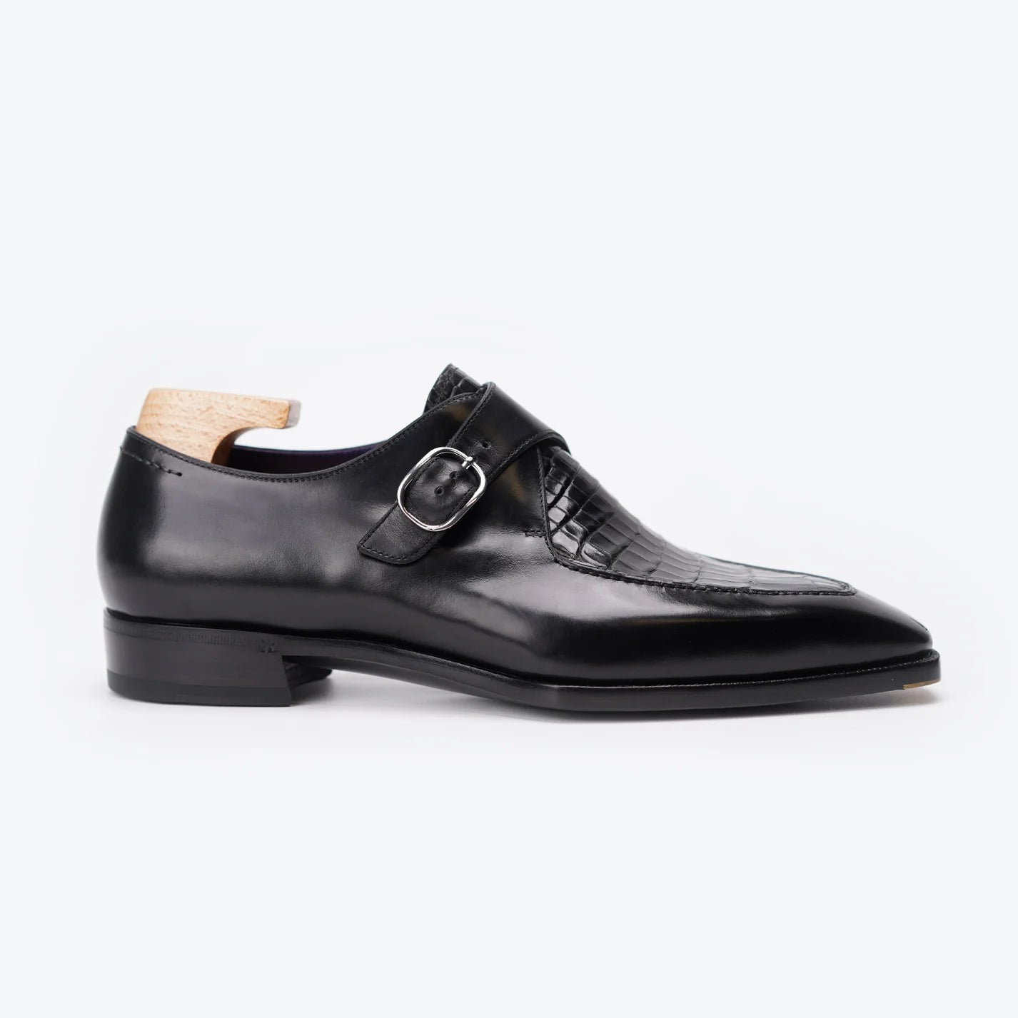 A black double monk strap leather shoe with a toe cap featuring a crocodile-embossed pattern, displayed on a light wood shoe tree against a clean white background.