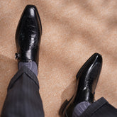 A pair of men's black leather dress shoes with crocodile-embossed panels on the toes, styled with dark pinstripe trousers and gray striped socks on a beige carpeted floor.