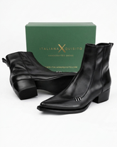 A pair of black leather ankle boots with low block heels and silver decorative studs, displayed in front of a green Italian X Quisito brand box on a white background.