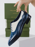 A navy blue leather wingtip oxford shoe with a white vachetta panel and brogue detailing, held by a hand in front of a green Italian Alps brand box.