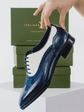 A navy blue leather wingtip oxford shoe with a white vachetta panel and brogue detailing, held by a hand in front of a green Italian Alps brand box.