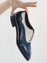 A hand holds a navy blue and white leather wingtip oxford shoe against a clean white background.