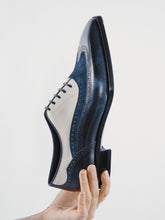 A hand holds a men's wingtip oxford shoe against a plain light gray background. The shoe features a dark navy blue leather upper with a contrasting white leather side panel and matching navy laces.