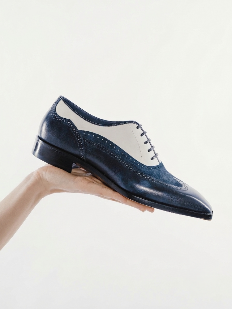 A navy blue leather wingtip oxford shoe with white leather panels, held by a hand against a clean white background.