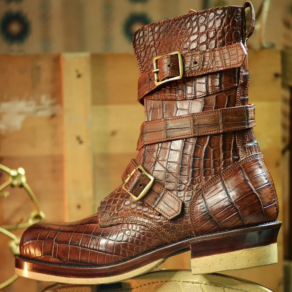 A brown embossed leather ankle boot with multiple buckle straps, shown standing on its beige sole against a rustic wooden background.