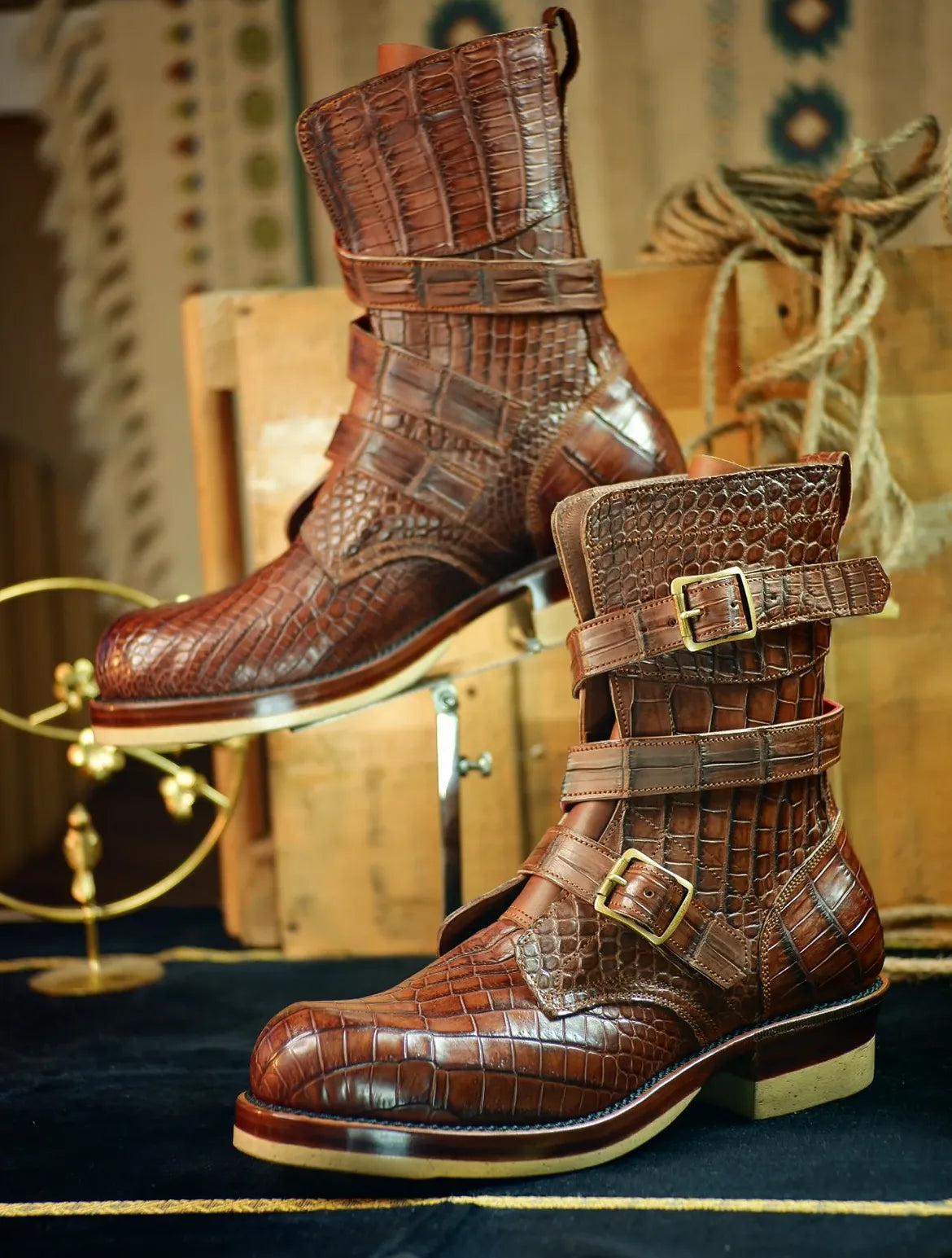 A pair of brown embossed crocodile leather ankle boots with buckle straps, displayed on a dark blue surface with vintage wooden crates and rope in the background.