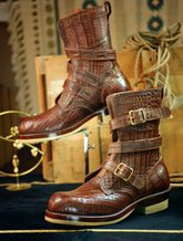 A pair of brown embossed crocodile leather ankle boots with buckle straps, displayed on a dark blue surface with vintage wooden crates and rope in the background.