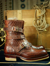 A pair of brown crocodile-embossed leather ankle boots with double buckle straps, displayed on a dark surface with a rustic wooden crate and rope in the background.