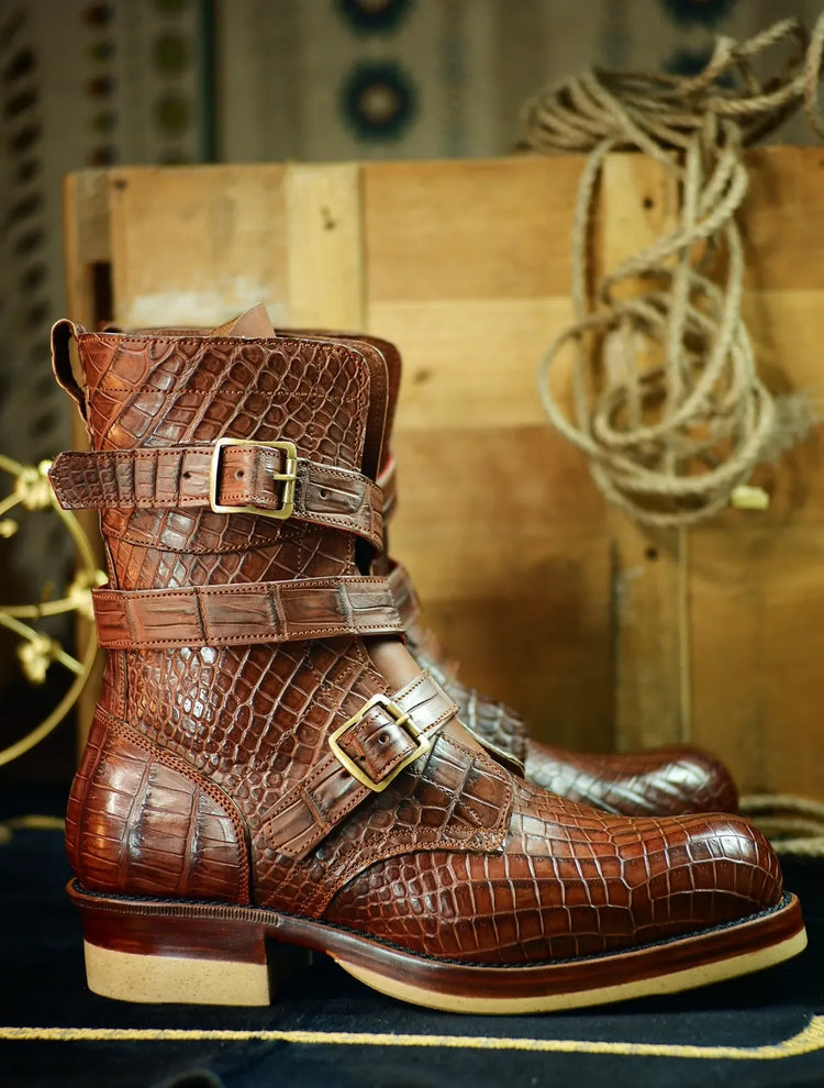 A pair of brown crocodile-embossed leather ankle boots with double buckle straps, displayed on a dark surface with a rustic wooden crate and rope in the background.
