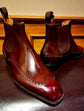 A pair of dark reddish-brown leather Chelsea boots with brogue detailing and elastic side panels, displayed on a dark surface with a wooden background.
