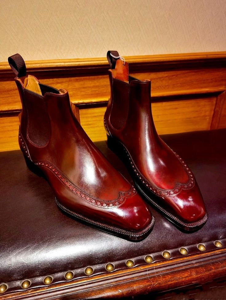 A pair of glossy dark red-brown leather Chelsea boots with brogue detailing, displayed on a dark brown leather bench with brass nailhead trim against a wood-paneled wall.