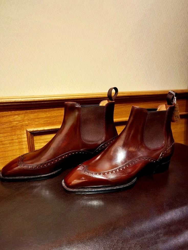 A pair of dark brown leather Chelsea boots with brogue detailing, displayed on a dark brown leather surface against a beige wall and wooden panel background.