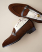 A pair of brown suede penny loafers with cream-colored insoles, white laces, and dark brown leather straps with silver-tone buckles, displayed on a neutral beige background.