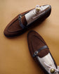 A pair of brown suede loafers with dark brown leather straps and silver buckles, displayed on white shoe trees with white rope laces and gold button details, set against a warm tan background.