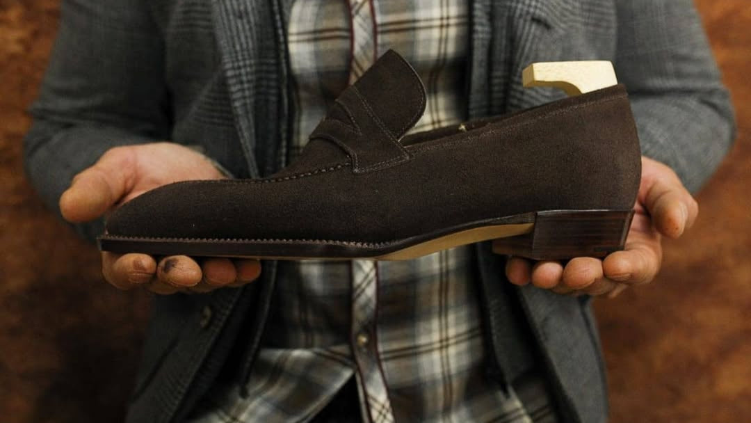 Person holding a brown suede loafer against a plaid shirt and gray jacket background