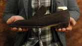 Person holding a brown suede loafer against a plaid shirt and gray jacket background