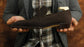 Person holding a brown suede loafer against a plaid shirt and gray jacket background