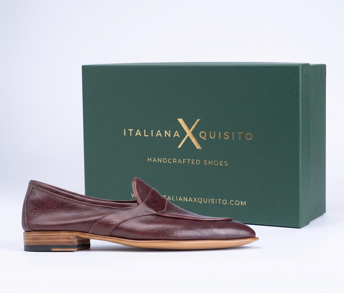 A single burgundy leather loafer shoe with a stacked wooden heel, positioned in front of its dark green branded shoebox on a white background.