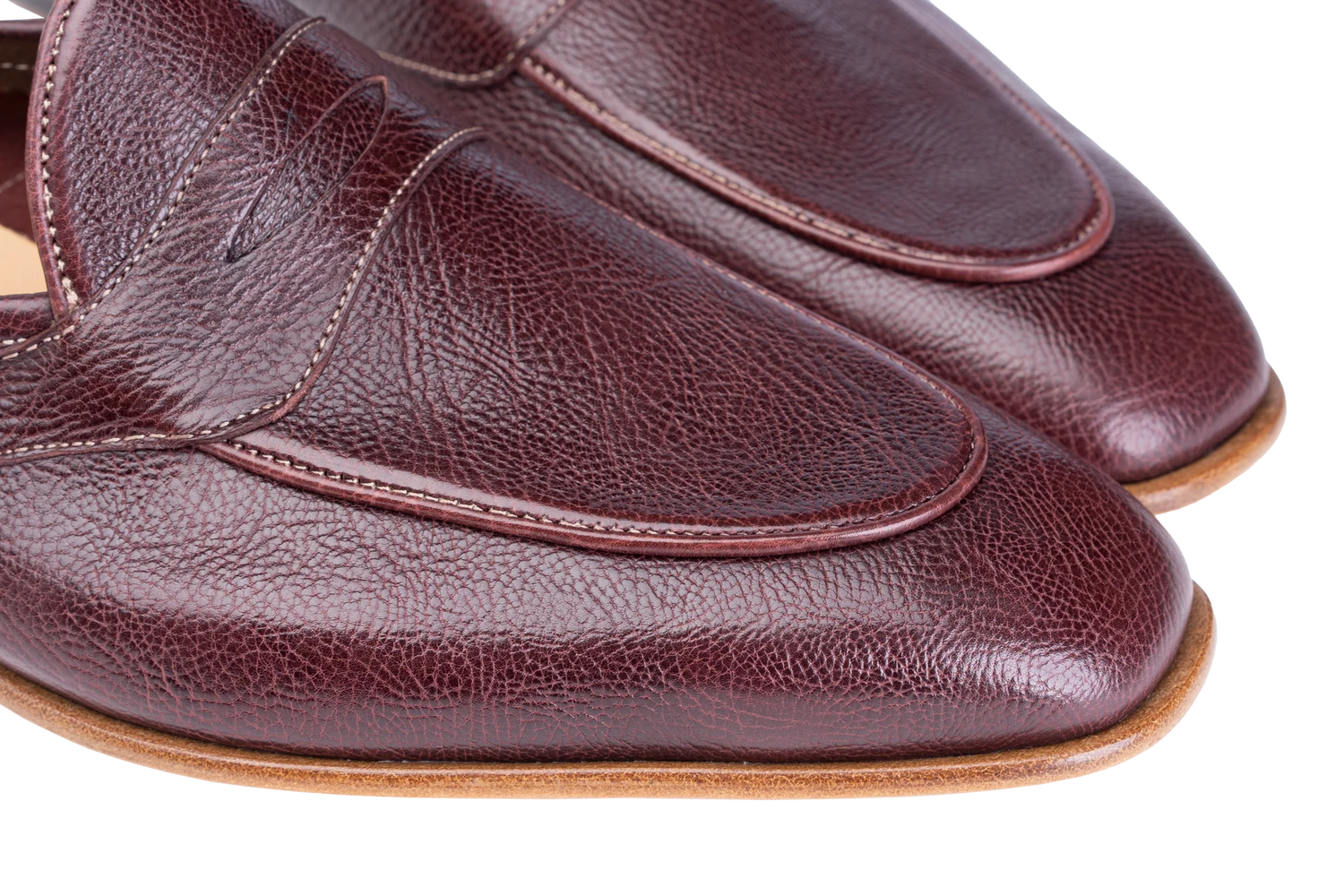 A close-up of a pair of men's penny loafers in dark burgundy pebbled leather, showing the toe, vamp, and side stitching against a white background.