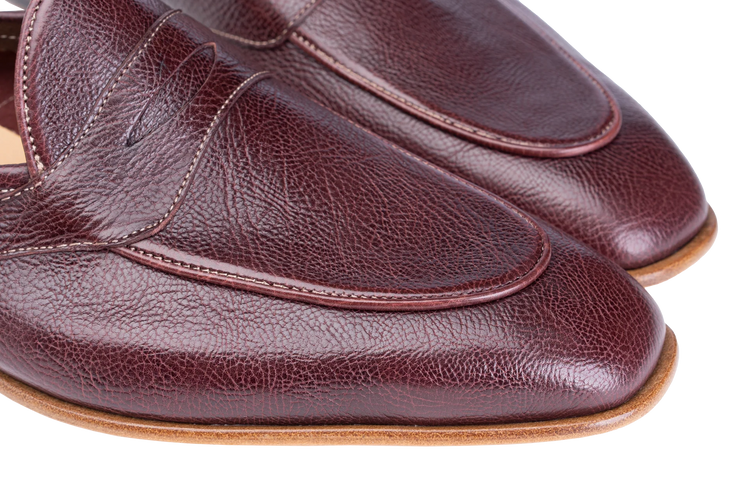 A close-up of a pair of men's penny loafers in dark burgundy pebbled leather, showing the toe, vamp, and side stitching against a white background.