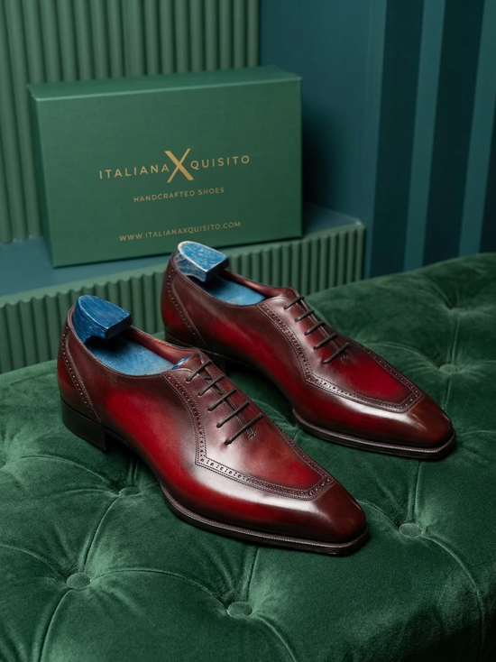 A pair of dark red brogue leather dress shoes with blue insoles, displayed on a green velvet tufted bench with the green Italian X Quisito shoebox in the background.