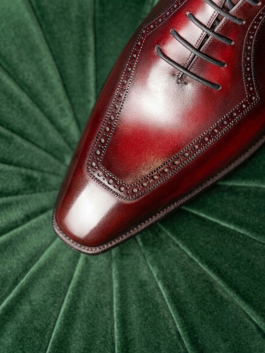 A close-up of a polished dark red oxford shoe toe with decorative brogue detailing, resting on a textured green velvet background.