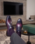 A pair of men's brogue dress shoes with a glossy purple and blue gradient finish, styled with dark purple socks and gray trousers. The shoes are shown with legs crossed in a modern living room setting next to a green box and a glass of whiskey.